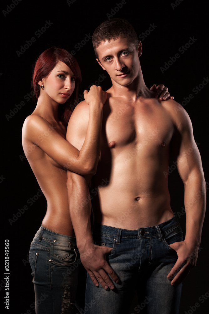 Front view of beautiful young couple posing topless. Attractive lady wrapping arm around shoulders of handsome man with naked torso. Isolated on black studio background. Concept of love and sexiness.