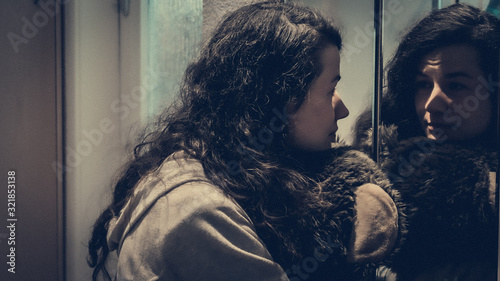 A woman starring at her reflection, thinking, while hugging a teddy bear. Gloomy feelings with a pinch of hope.