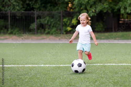 Toddler girl playing football: blonde child in white shirt and pink sneakers running on green soccer field to kick ball. Young sportsman, female power and active childhood concept. Copy space