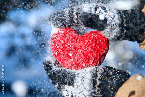 Valentine's day background.Hands in fluffy mittens hold a red knitted heart of thread on a background of a snowy forest.Close-up, cropped shot, horizontal.Concept of a holiday and relationship.