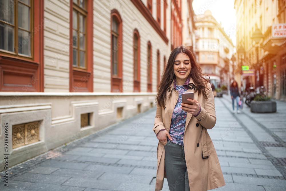 Fototapeta premium Gorgeous beautiful young woman with brown hair messaging on the smart-phone at the city street background. pretty girl having smart phone conversation in sun flare.