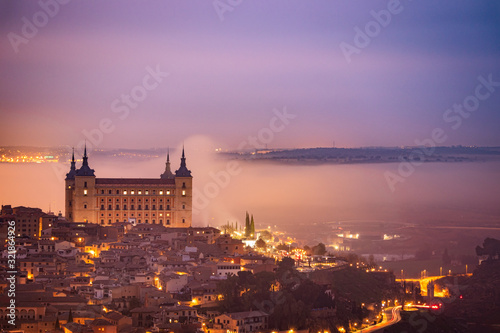 Fototapeta Naklejka Na Ścianę i Meble -  Sunrise in the historic and amazing Downtown Toledo. Alcazar is surrounded by fog. Foggy morning with city lights in Toledo, Spain.