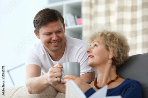 Elderly woman happily takes care from an adult son. An elderly woman in casual clothes sits at home on couch and holds in her hands cup that man gives her. Son expresses warm feelings mother.