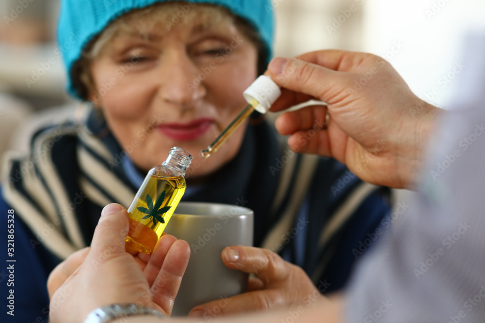 Fototapeta premium Elderly woman receives sedative drops from doctor. Close up an elderly woman looking at cup. Doctor collected carly hemp oil and drips them into patient cup. Prescription herbal drops.