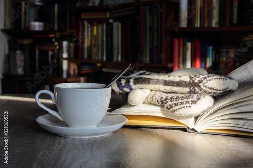 A white cup with coffee stands on a table lit by the sun. Opposite shelves with books. Nearby is an open book with mittens. Concept - writing, coffee break, winter evening