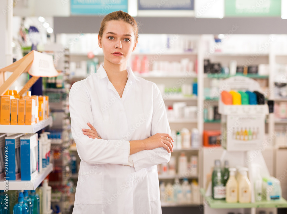 Cheerful woman pharmacist standing with arms crossed in interior of pharmacy