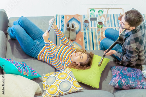 Boy singing with a guitar and a girl making a selfie with a tablet. Young couple in the sofa, relax moment. Relax, love and technology concept. Focus on girl face - Image