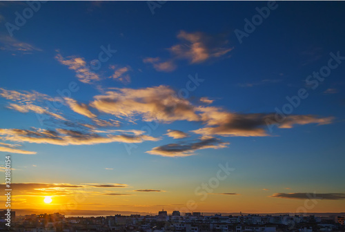 Scenic cityscape with golden evening sun goes down over the horizon. Gold colored sunset clouds in a clear blue sky.