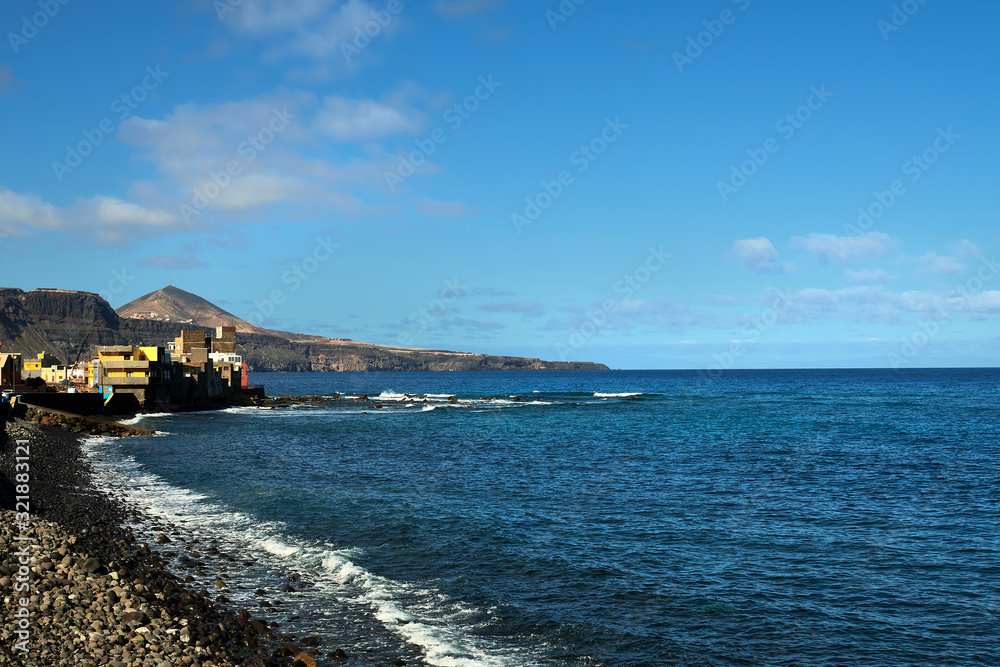 Coast with some houses and mountains in the background. Calm sea and blue sky with some clouds. Coast with some houses and mountains in the background. Calm sea and blue sky with some clouds.