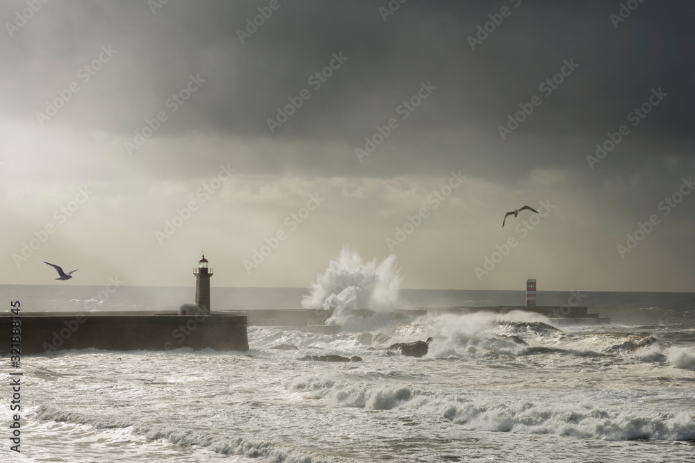 Great marine wave breaking over the breakwater of the Foz do Douro lighthouse in Porto, seagulls, sun rays, marine storm under a cloudy sky