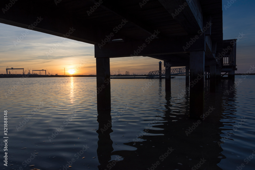 coastal landscape with wooden jetty reflected in calm water under a sunset with clouds and large industrial cranes in the background