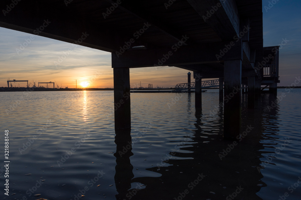 coastal landscape with wooden jetty reflected in calm water under a sunset with clouds and large industrial cranes in the background coastal landscape with wooden jetty reflected in calm water under a sunset with clouds and large industrial cranes in the background