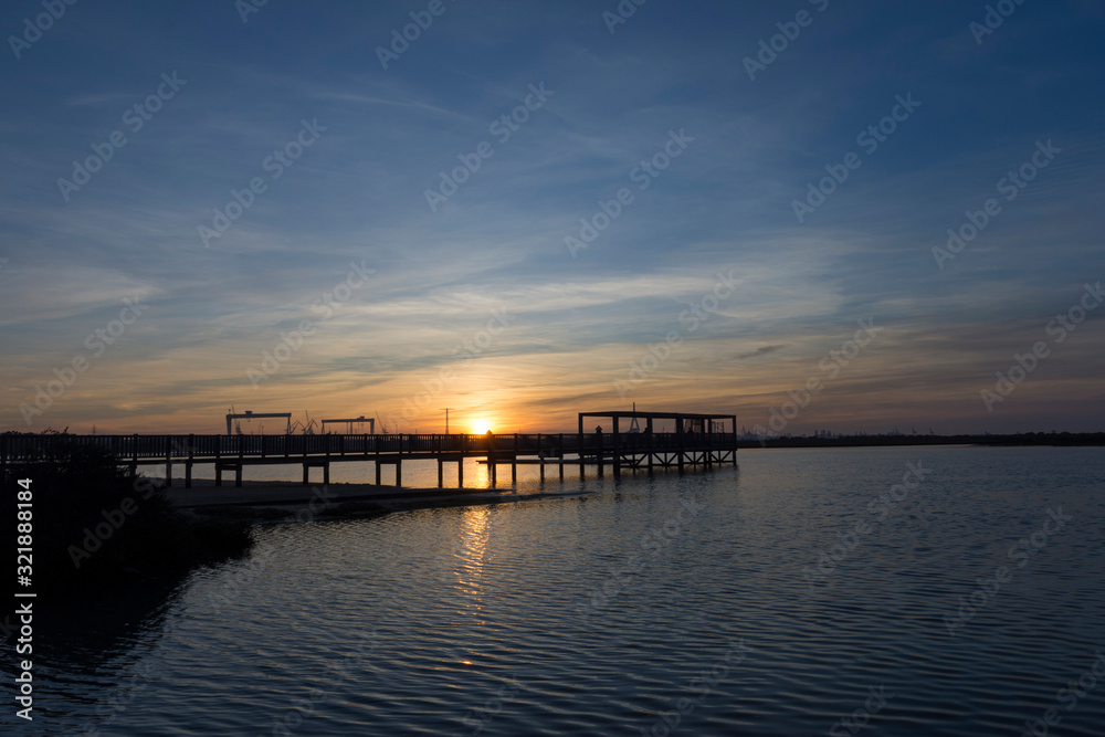 coastal landscape with wooden jetty reflected in calm waters under a sunset with clouds and large industrial cranes in the background