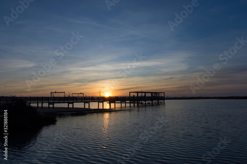 coastal landscape with wooden jetty reflected in calm waters under a sunset with clouds and large industrial cranes in the background © roferfrann