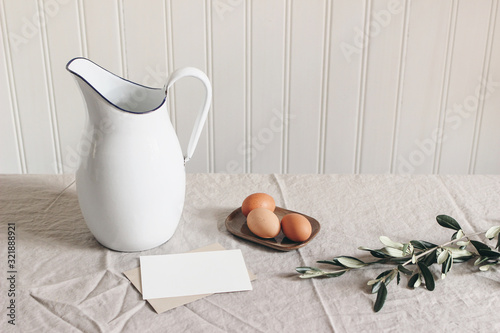 Spring breakfast still life scene. White jug, olive branch, chicken eggs on linen tablecloth. Blank greeting card mockup. Easter food and drink concept. Farmhouse table composition. Scandi interior. photo