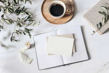 Summer wedding stationery scene. Open diary and blank greeting card mockups. Cup of coffee wooden plate, book, ribbon, olive leaves and branches on white marble table background. Flat lay, top view.