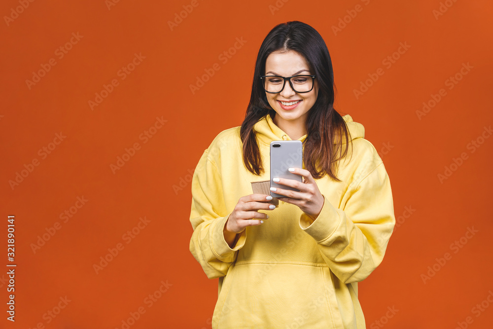 Portrait of a pretty young woman using mobile phone while holding takeaway coffee cup isolated over orange background. Portrait of a pretty young woman using mobile phone while holding takeaway coffee cup isolated over orange background.