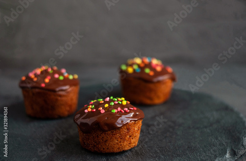 Pumpkin muffins are poured with chocolate ganache, decorated with colored confectionery on a black concrete background.