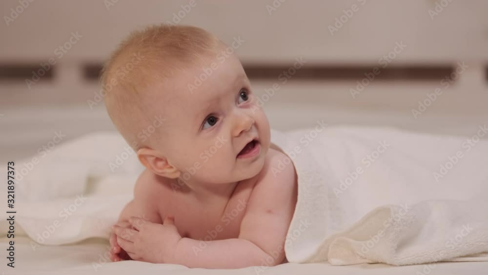 Smiling little baby girl with big blue eyes lying on her tummy on white bed looking at camera and smiling. happy baby in white lies on the bed and touches the legs with his hands. Smiling little baby girl with big blue eyes lying on her tummy on white bed looking at camera and smiling. happy baby in white lies on the bed and touches the legs with his hands.