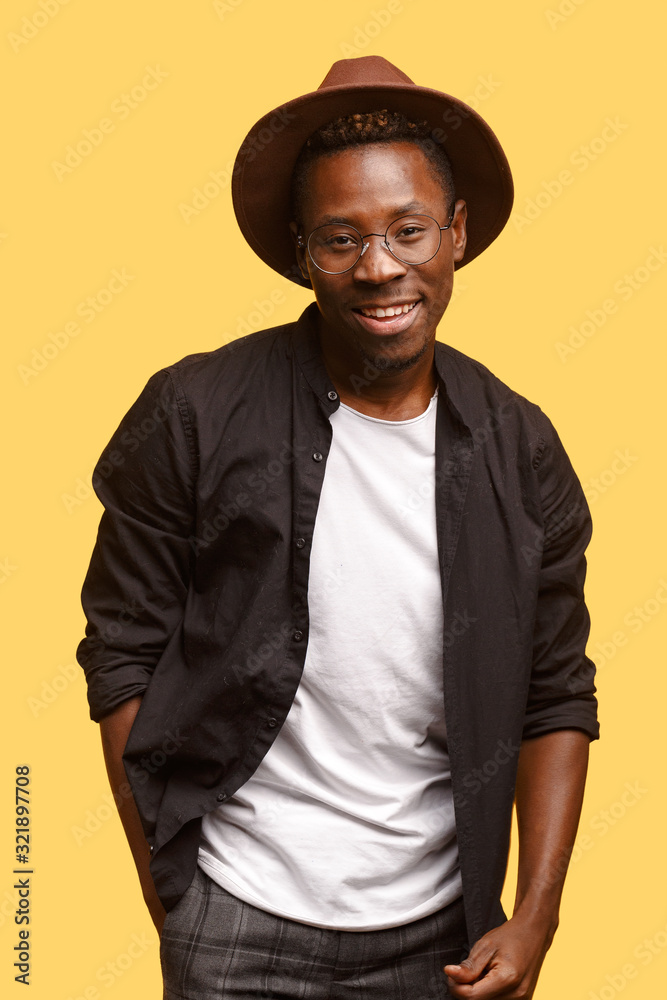 Hilarious African guy in hat, glasses and black shirt poses on a yellow background. Studio shooting.
