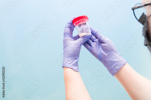 a woman in blue disposable gloves holds a jar of sperm, a red cap, a blue background, a woman with glasses and a black medical mask, copy space, donate sperm for analysis, sperm donor concept