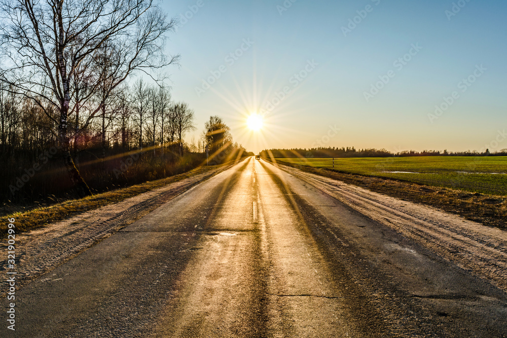 The asphalt of an old road, cracked in the cold, is lit by the sun's rays in a haze of blue sky against the background of a overed fields and trees