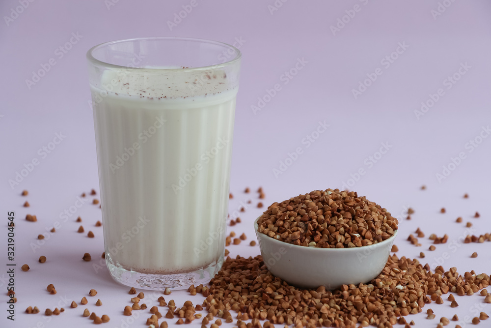 Glass beaker with milk on lilac background. Next to the glass is a bowl of buckwheat, part of which is scattered on the table. Plant based milk concept. Glass beaker with milk on lilac background. Next to the glass is a bowl of buckwheat, part of which is scattered on the table. Plant based milk concept.
