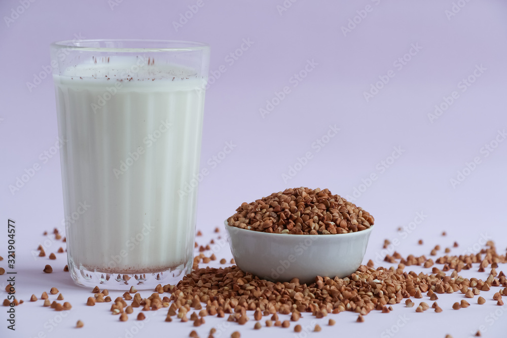 Glass beaker with milk on lilac background. Next to the glass is a bowl of buckwheat, part of which is scattered on the table. Plant based milk concept. Glass beaker with milk on lilac background. Next to the glass is a bowl of buckwheat, part of which is scattered on the table. Plant based milk concept.