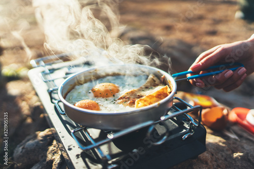 person cooking fried eggs in nature camping outdoor, cooker prepare scrambled breakfast picnic on metal gas stove, tourism recreation outside; campsite lifestyle