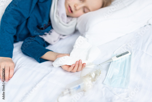 Caucasian 7 year old sick boy sleeping with handkerchief in hand and protective mask, thermometer on the bed. Virus, contagious disease concept.