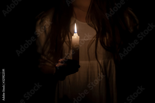 Photo of a candle at night holding by a young girl in an old white dress.  Focus on the candle. Dark background. Scary horror concept. 