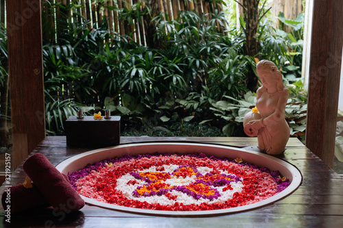 Outdoor jungle view stone round bath tub with flower shaped petals in pink,red,white colors near window. Organic spa relaxation in luxury Bali bathroom.