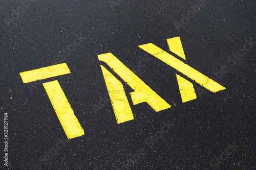 A bright yellow word TAX painted on an asphalt car parking surface © Chris