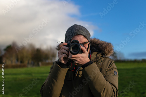 Man taking photos with digital camera in front, dressed in a coat and hat on a beautiful meadow with grass and trees on a sunny day.