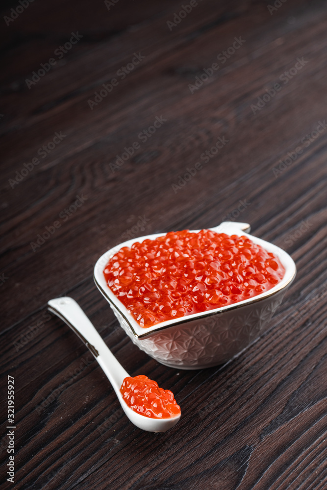 Red caviar in a white ceramic bowl with spoon on a brown rustic wooden background Red caviar in a white ceramic bowl with spoon on a brown rustic wooden background