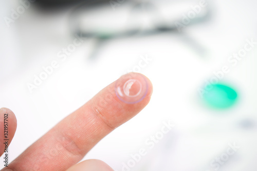 Hand with contact lens prepared to insert into eye. Glasses  drops  and cases visible in the background