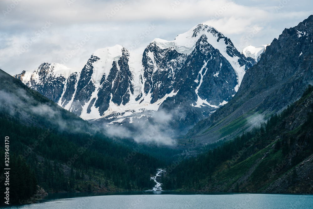 Beautiful big glacier, rocky snowy mountains, coniferous forest on hills, mountain lake and highland creek under cloudy sky. Atmospheric alpine scenery with low clouds on high forest steep slopes.