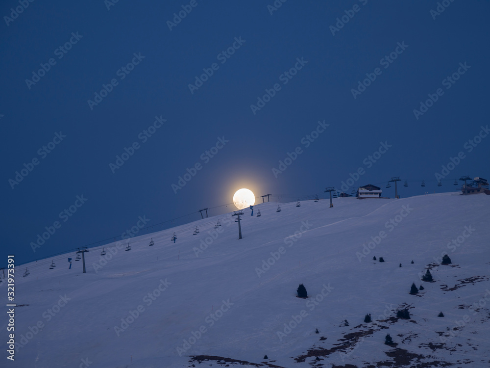 Ski slopes illuminated by the light of a giant moon. The moon rises from the top of the mountain. Monte Pora ski resort. Orobie. Italian Alps. Winter time