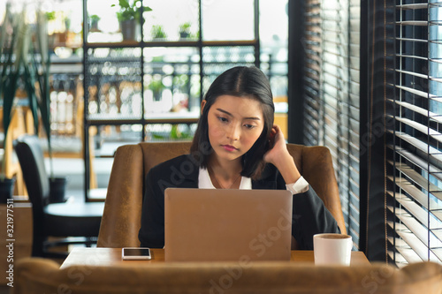 Close up charming young Asian woman in black suit sitting and working with laptop on wooden table with cell phone and coffee cup