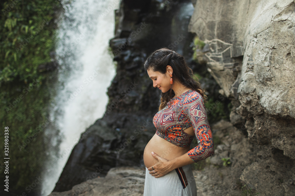 Portrait of young authentic pregnant woman near waterfall outdoors. Smiling and happy, holding belly. Spiritual harmony with nature. Travel and childbirth in Bali.