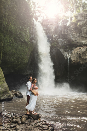Young pregnant couple in love with amazing view of Tegenungan cascade waterfall. Morning sunlight  Happy together  pregnancy travel lifestyle. Honeymoon in Bali.