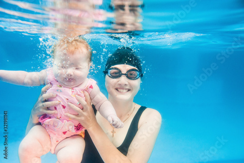 Young mother, swimming instructor and happy little girl in the pool swimming under water. Teaches infant child to swim. Diving baby in the paddling pool. Mother holding baby and dive. Close up.