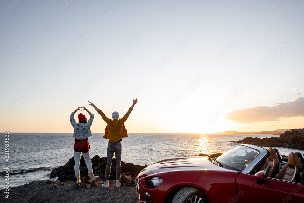 Fototapeta premium Couple enjoying beautiful views on the ocean, standing together with raised hands near the car on the rocky coast, wide view from the side with copy space on the sky