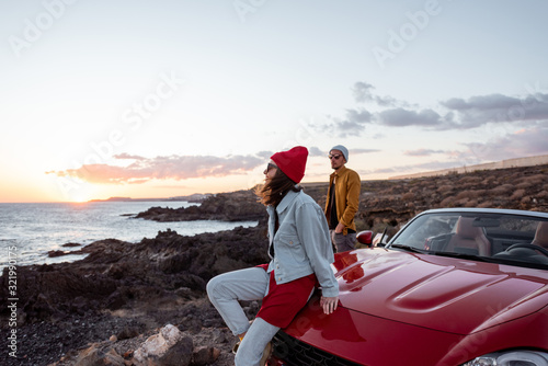 Young joyful couple having fun on the rocky coast while traveling by car on a sunset. Carefree lifestyle, love and travel concept