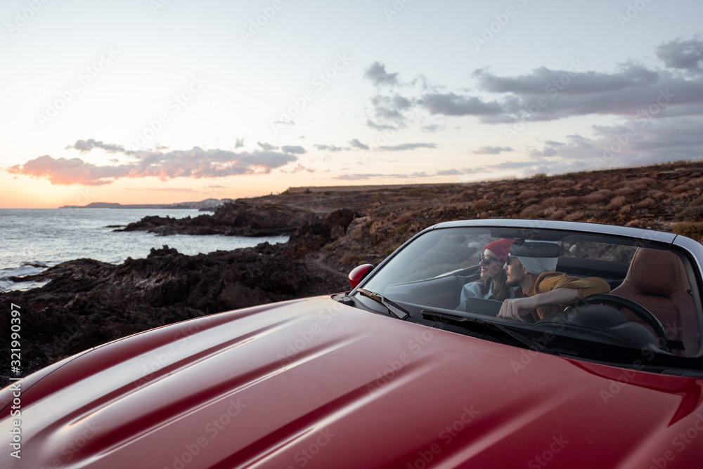 Couple driving convertible car, traveling near the ocean on a sunset, wide landscape view. Happy vacations and traveling by car concept