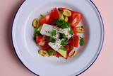 Traditional Greek salad with feta cheese and vegetables served in white plate over bright pastel pink background.