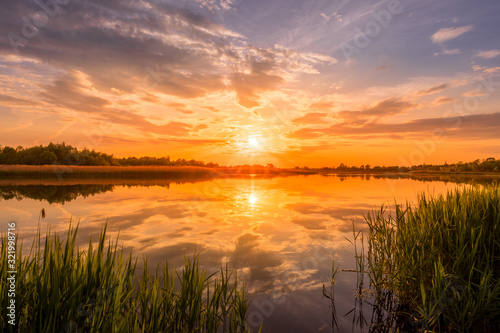 Fototapeta Naklejka Na Ścianę i Meble -  Scenic view of beautiful sunrise or dawn above the pond or lake at spring or early summer morning with cloudy sky background, fog over water and reed grass with dew at foreground. Landscape. Water ref