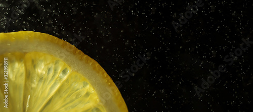 Juicy slice of lemon on a dark background. In the frame, only part of the citrus. Long banner. Drops of water in the air. Detailed macro photo. Copyspace, minimalism.