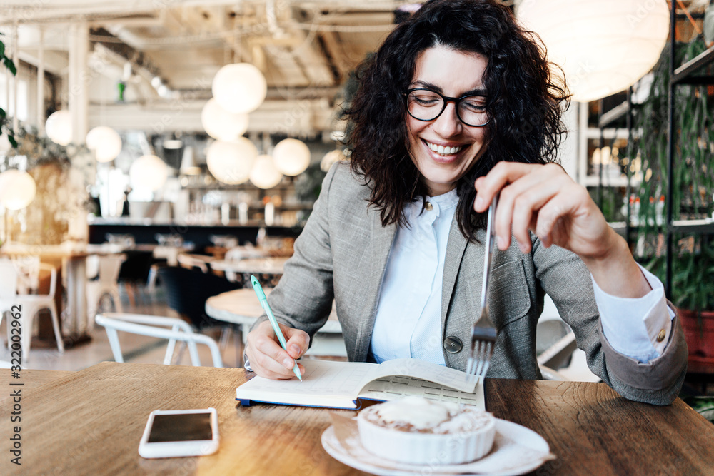 Young curly woman laughs and eats sweet dessert in coffee house. Working with note book and mobile phone