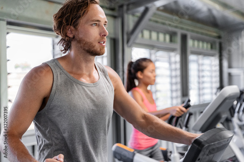 Fitness gym people training on elliptical cardio machine trainer. Interracial couple friends, Caucasian man, Asian woman working out together at gym health center.